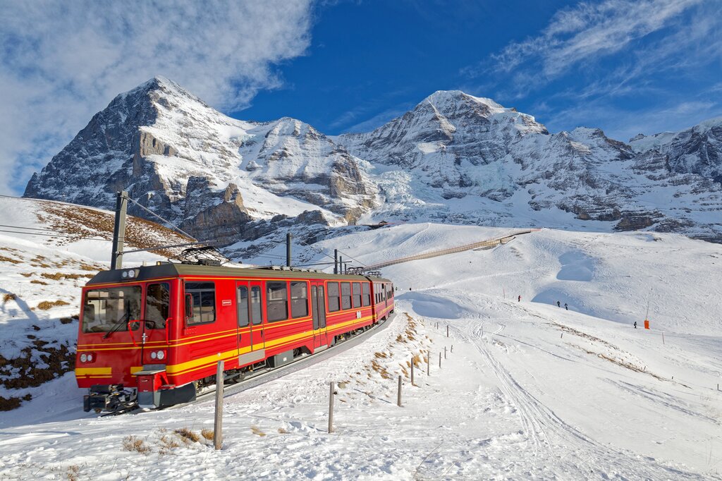 On a sunny winter day, tourists ride on a cogwheel train from Jungfraujoch (Top of Europe) to Kleine Scheidegg on the snowy hillside with Eiger and Monch in background, in Berner Oberland, Switzerland